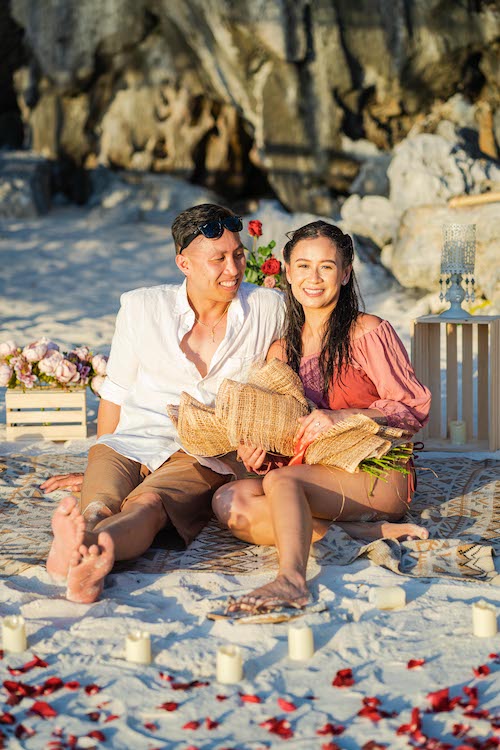 A couple sits on a beach surrounded by candles and petals, setting the stage for a picturesque proposal. The man, in his white shirt and sunglasses, smiles at the woman holding a bouquet and wearing a pink blouse. They sit on a patterned blanket with a stone cliff in the background.