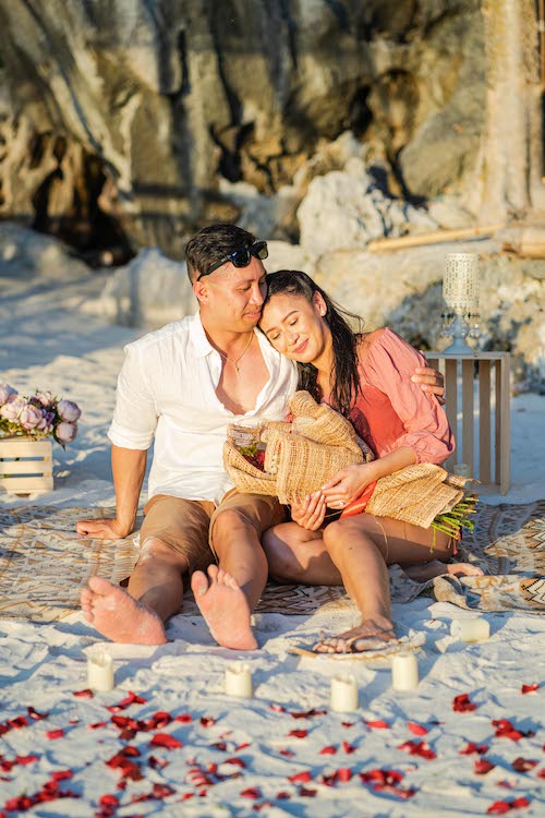 A couple sits on a beach, surrounded by candles and flower petals. The man, in a white shirt and shorts, embraces the woman in a pink dress holding a bouquet. This picturesque setting whispers of an intimate proposal as they appear relaxed and joyful in this natural paradise.