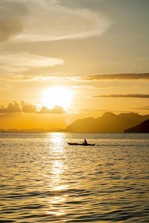 During a serene tour, a person kayaks on calm waters as the golden sunset illuminates the scene. The sun sets over distant mountains, casting a warm glow over the sky and water, with clouds reflecting the sunlight. It's an ideal backdrop for a perfect proposal.