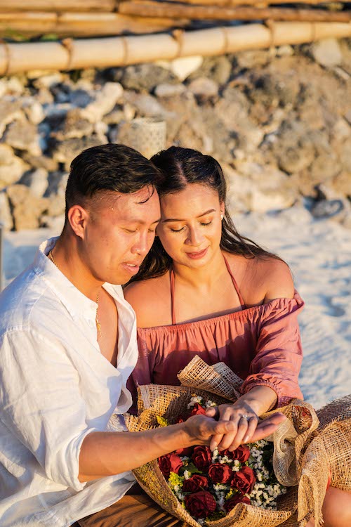 A couple sits on a sandy beach at sunset, the perfect setting for a marriage proposal, holding hands and a bouquet of roses. Dressed casually, with the man in a white shirt and the woman in an off-shoulder pink top, they are surrounded by rocks and a serene atmosphere.