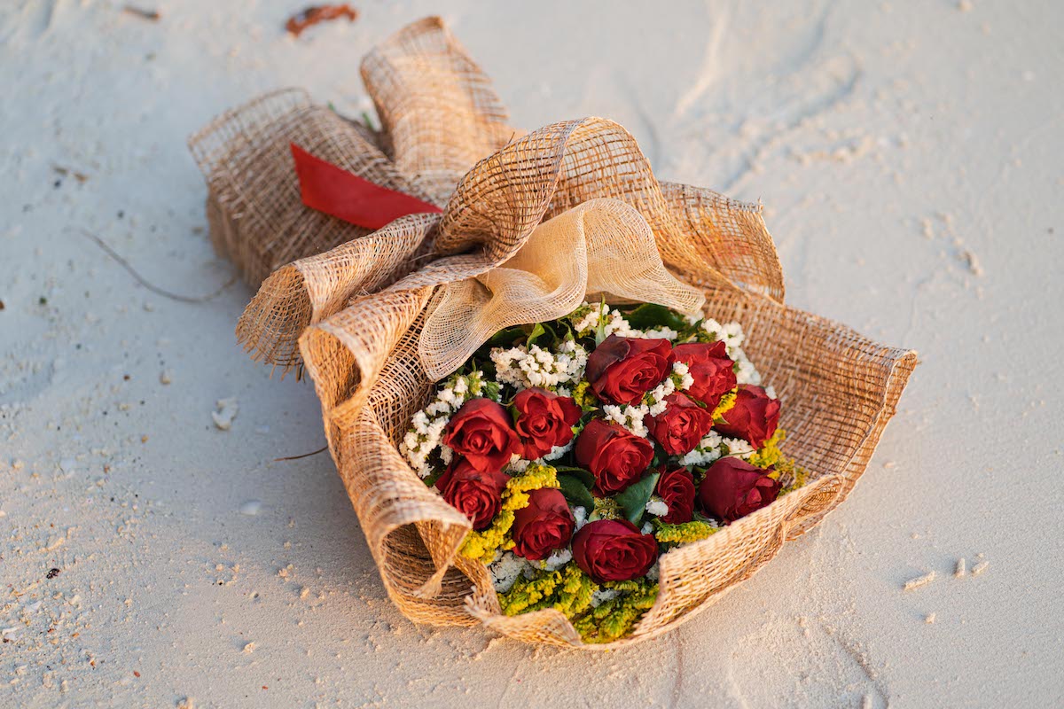A bouquet of red roses wrapped in burlap with a red ribbon, perfect for a marriage proposal, rests on a sandy surface. White and yellow filler flowers mix with the roses, adding texture and contrast to the arrangement.