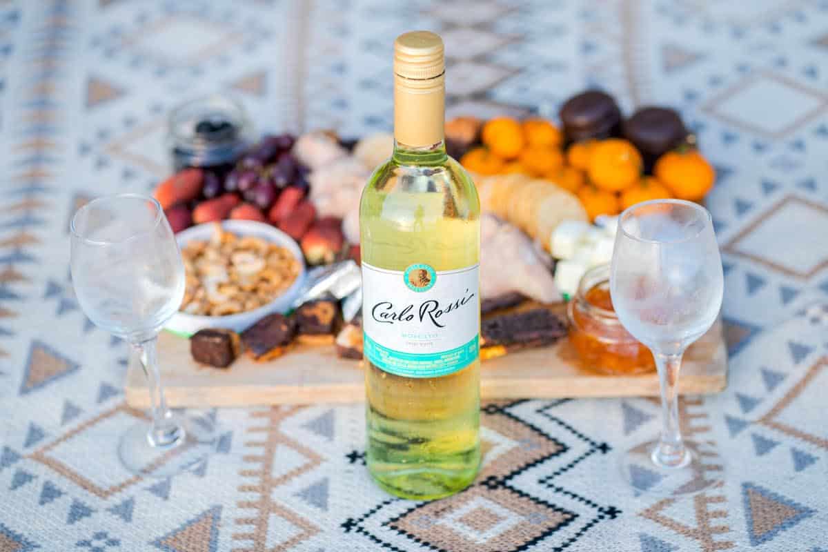 A bottle of Carlo Rossi white wine sits on a patterned picnic blanket with two empty wine glasses waiting for a toast. Behind it, a charcuterie board holds a variety of snacks, including fruits, crackers, and cheeses—a perfect setting for a romantic proposal.