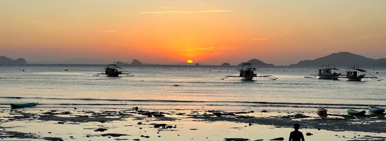 Screenshot A serene sunset over a calm sea in Palawan reveals silhouettes of traditional boats and distant deserted islands. The sky, a gradient of orange and yellow, reflects on the water. A person stands on the beach, framed by scattered seaweed and small boats, dreaming of a tranquil boat tour.