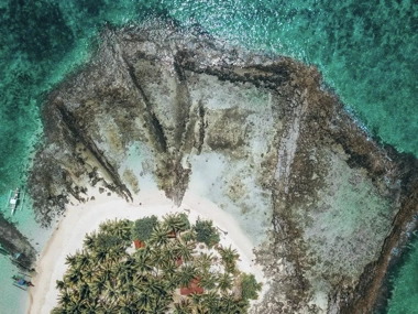 Aerial view of a small, palm-covered island surrounded by clear turquoise water and rocky formations, perfect for island hopping on a scenic boat tour.