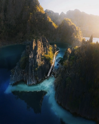 Aerial view of a narrow bridge connecting jagged limestone islands, perfect for island hopping, surrounded by deep blue water. Trees and sunlight illuminate the scene, inviting a serene boat tour experience.