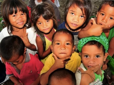 A group of smiling children pose playfully, some with hands on their chins, looking up at the camera, excited after an adventurous boat tour. They are outdoors with trees in the background, making it a perfect day for an island hopping trip.