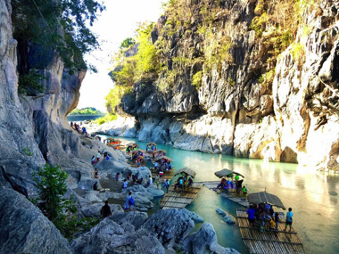 Bamboo rafts float on a clear river flanked by rocky cliffs, with people enjoying a boat tour along the riverbank under a bright sky.