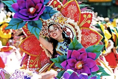 A dancer in vibrant traditional costume with colorful floral headdress and decorations performs joyfully at the parade, capturing the spirit of island hopping adventures.