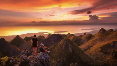 Two people admire a vibrant sunset from a mountaintop, overlooking rugged hills and a distant body of water where a boat tour awaits to whisk them away for some island hopping adventures.