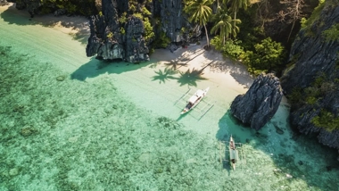 Aerial view of a tropical beach with clear turquoise water, rock formations, palm trees, and a small boat poised for an island-hopping adventure along the pristine shoreline.
