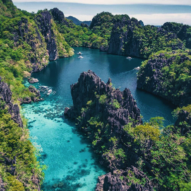 Aerial view of a vibrant turquoise lagoon, perfect for island hopping or a boat tour, surrounded by lush, jagged cliffs and forests under a clear blue sky.