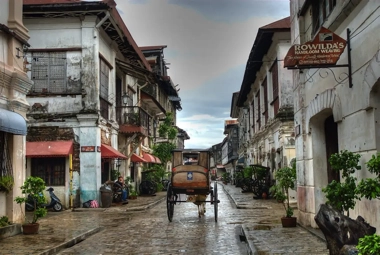 A narrow, cobblestone street lined with old buildings, featuring a horse-drawn carriage meandering along, evokes the charm of island hopping adventures, as tendrils of greenery sway gently on a cloudy day.