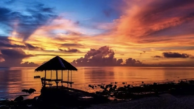 A tropical hut on a rocky beach at sunset, with vibrant orange, pink, and purple clouds reflecting on calm water. Nearby, island hopping adventures await those eager to explore the scenic wonders beyond the shore.
