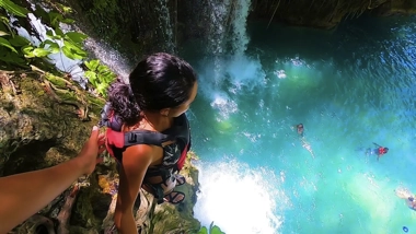 Standing at the edge of a tropical waterfall with a backpack, they gaze down at the blue water where swimmers enjoy the day, dreaming of their next adventure—perhaps island hopping or a boat tour—to explore even more hidden gems.