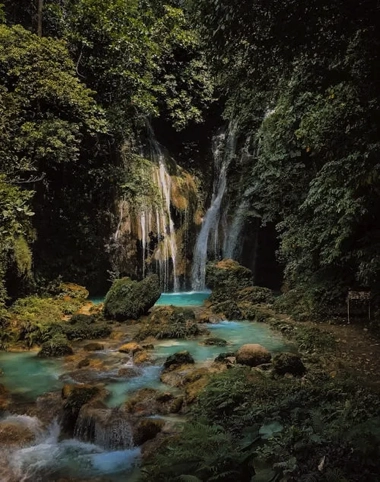 A serene waterfall cascades into a turquoise pool, inviting visitors from their boat tour to revel in its beauty, surrounded by lush green foliage and rocks.