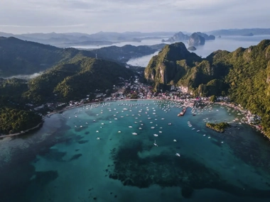 Aerial view of a bay surrounded by lush green mountains, perfect for island hopping, dotted with numerous boats offering boat tours on the clear blue water. Coastal town visible along the shore.