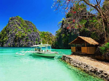 A small boat from a thrilling island hopping adventure floats on clear turquoise water near rocky cliffs and a bamboo hut, under a bright blue sky with a tree branch in the foreground.