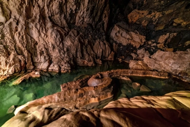The dimly lit cave features a small pool of greenish water surrounded by rocky formations and textured walls, making it an intriguing stop on your island-hopping adventure.
