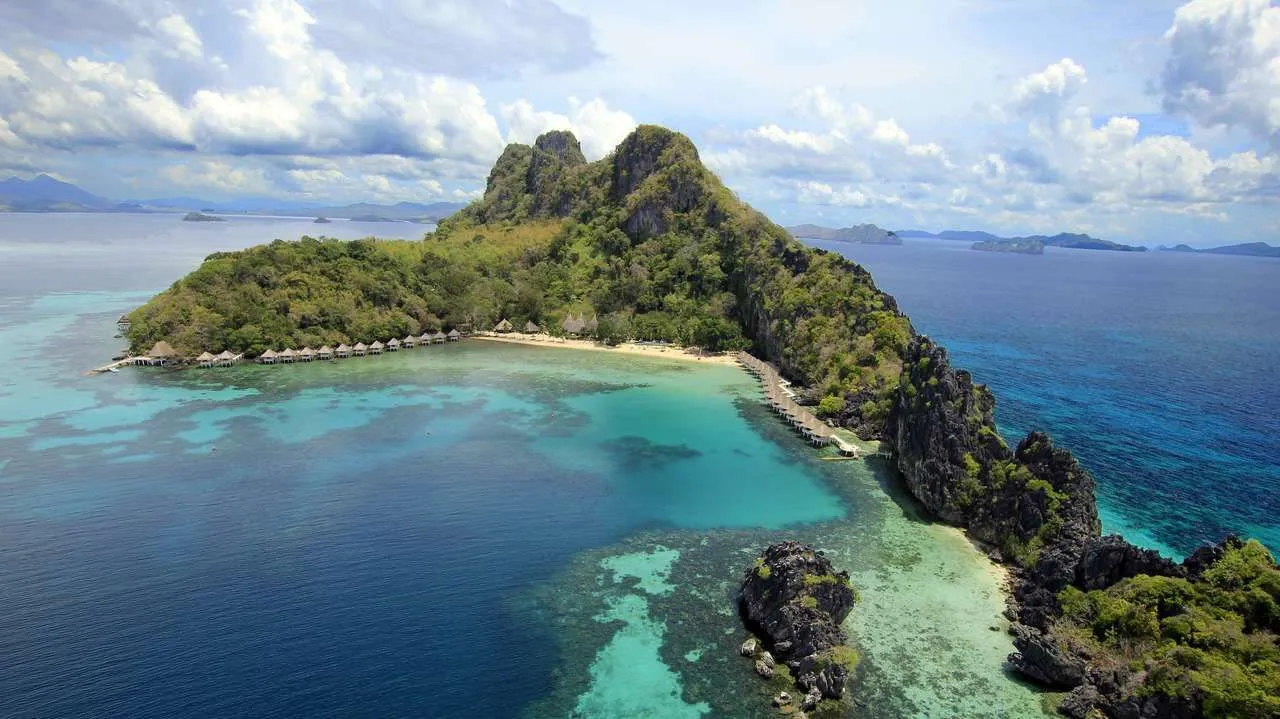 taytay bay sea turtles apulit island el nido resort Aerial view of a tropical island in Palawan with lush greenery and rocky cliffs, surrounded by clear turquoise and deep blue waters. Overwater bungalows line part of the shoreline while boat tours navigate below fluffy clouds dotting the sky above.