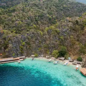 Aerial view of an exclusive island beach with vibrant turquoise waters, boats, and a lush forest backdrop.