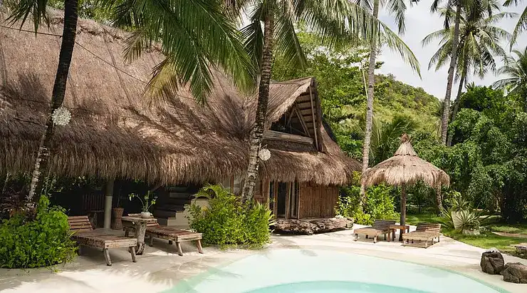Idyllic tropical island resort featuring traditional thatched-roof huts, outdoor lounge chairs, and lush tropical surroundings.