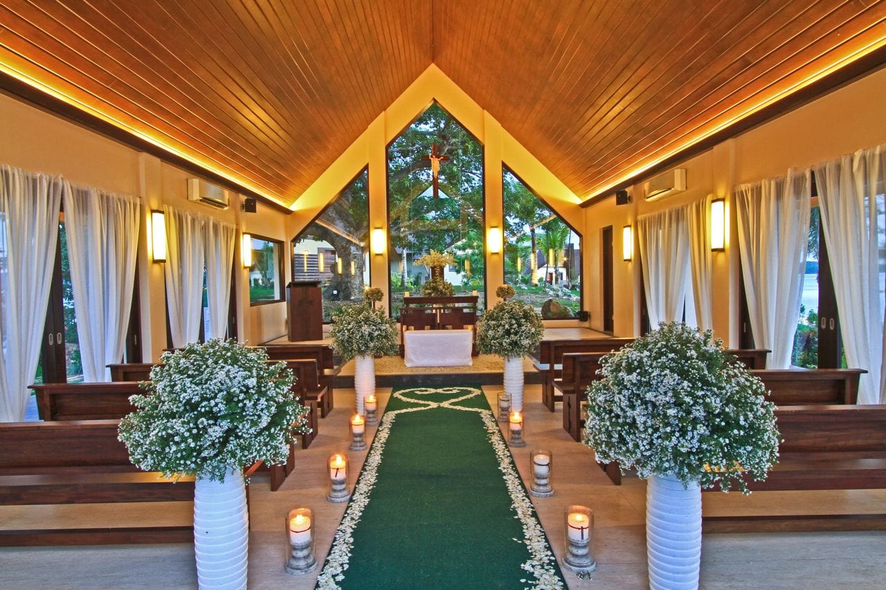 Chapel interior with wooden pews, white flowers, candles, and large windows.