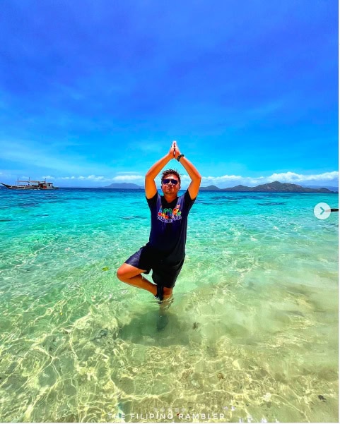 Person practicing yoga in shallow clear turquoise water on a tropical beach with boats and islands in the background.