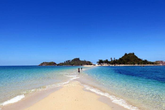 A sunny beach in Carles with clear blue water, a sandy path leading to the Gigantes Islands under a bright sky, and two people island hopping.