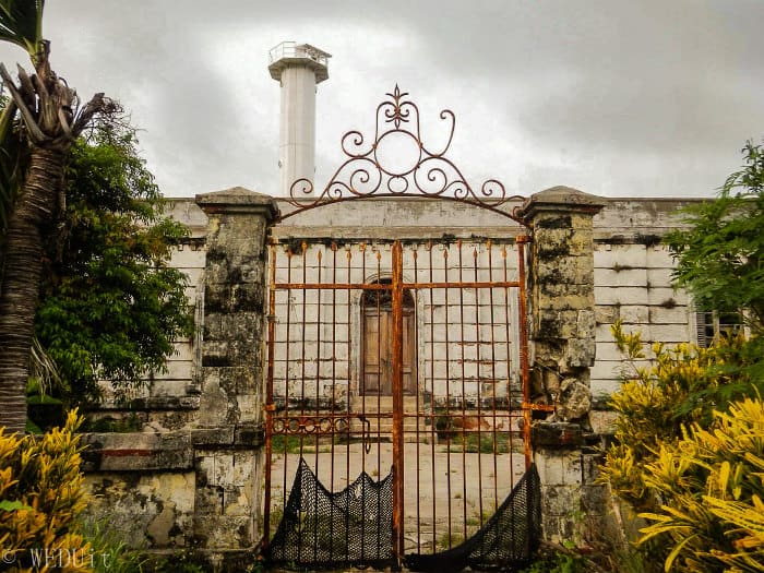 A rusty iron gate leads to an old stone lighthouse in Carles, with overcast skies and lush foliage, perfect for island hopping.