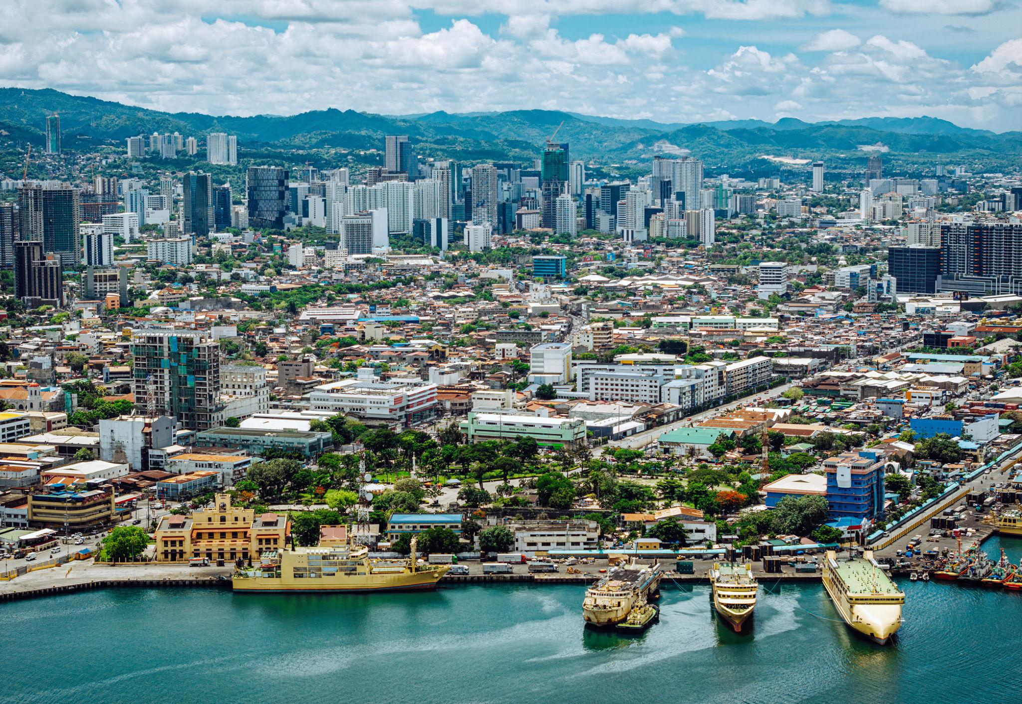 Aerial view of Cebu’s coastal city with tall buildings, mountains beyond, and ships docked along the vibrant waterfront.