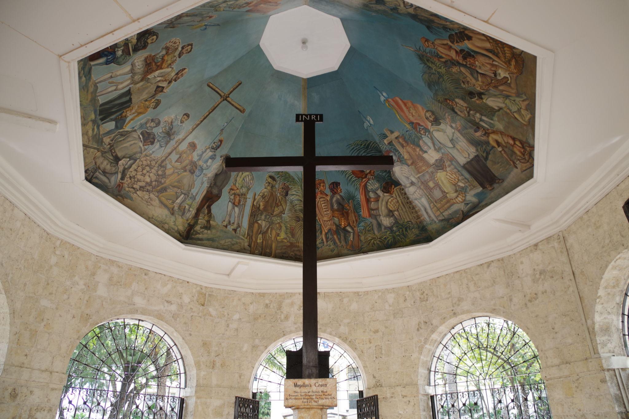 Large wooden cross under a painted dome depicting historical scenes, inside a Cebu stone gazebo with arched windows.