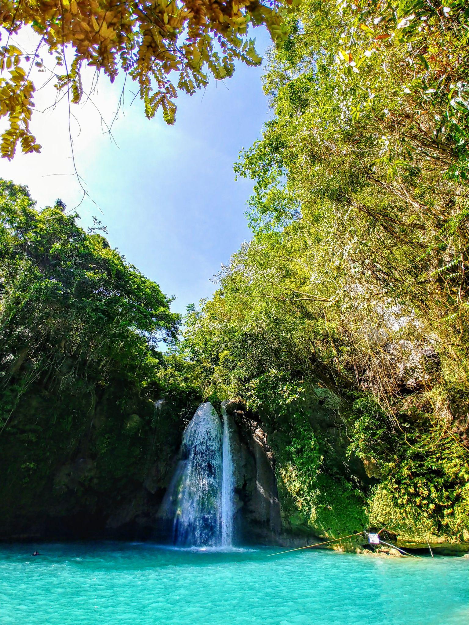 A vibrant waterfall cascades into a turquoise pool near Siquijor, surrounded by lush green trees under a clear blue sky.