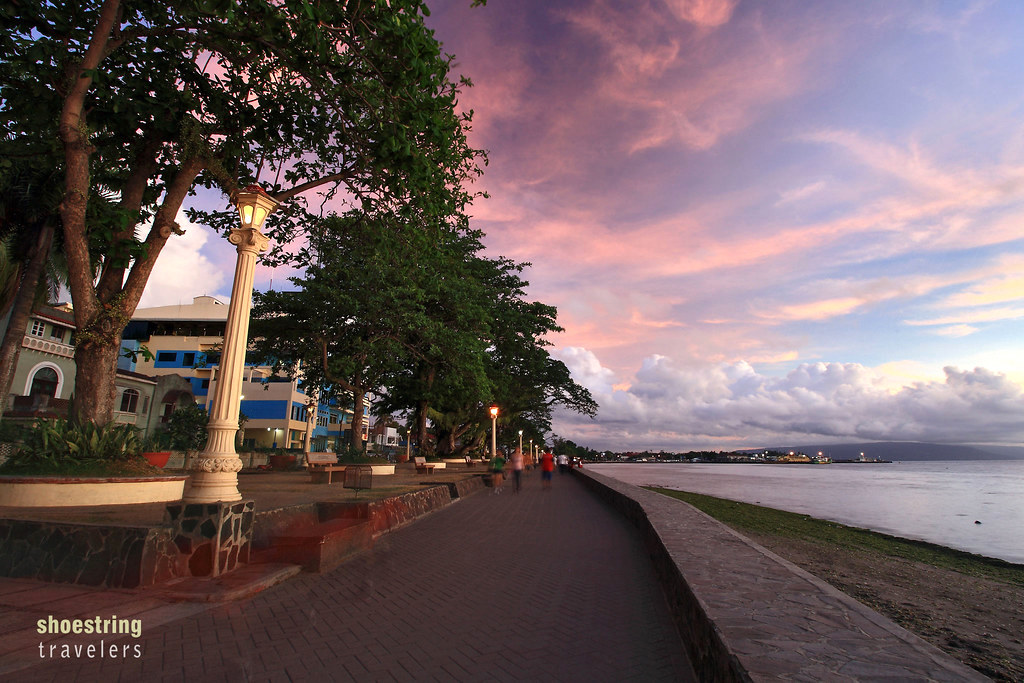 A scenic Dumaguete seaside promenade at sunset, with trees, lampposts, and calm water under a pink and purple sky.
