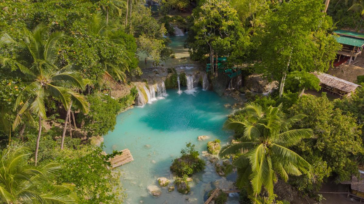 Aerial view of a turquoise waterfall pool in Cebu, surrounded by lush green trees, palm trees, and small wooden huts nearby.