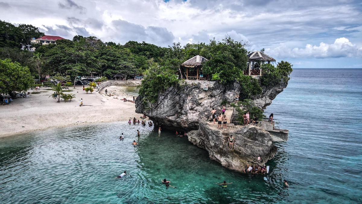 A rocky outcrop with huts overlooks a beach in Siquijor, where people swim and relax under a partly cloudy sky and clear blue water.