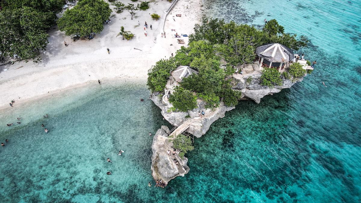 Aerial view of a tropical Siquijor beach with clear turquoise water, rocky outcrop, trees, and people swimming and relaxing on the sand.
