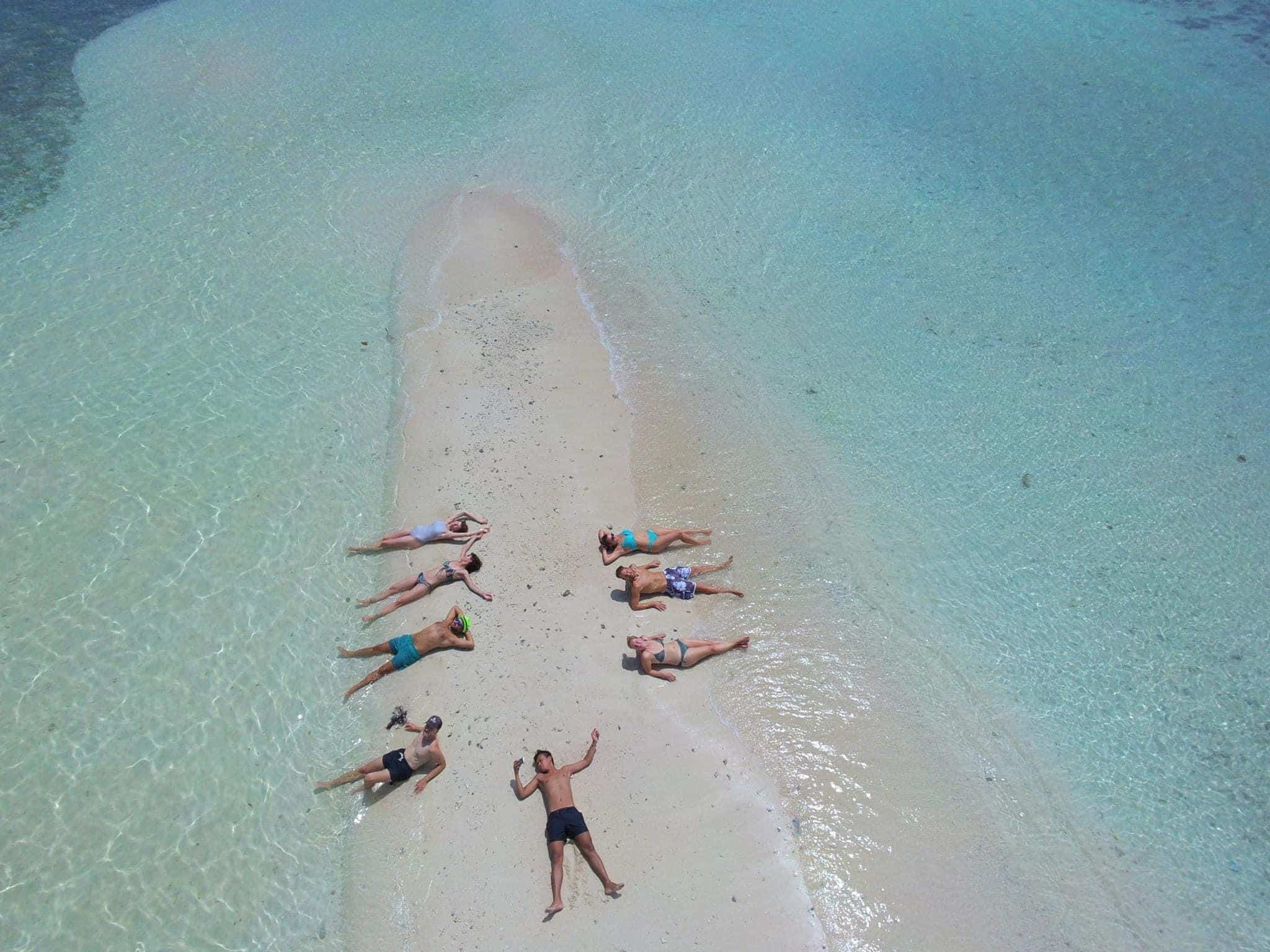 Aerial view of friends sunbathing and swimming in crystal-clear ocean waters near a sandy island.