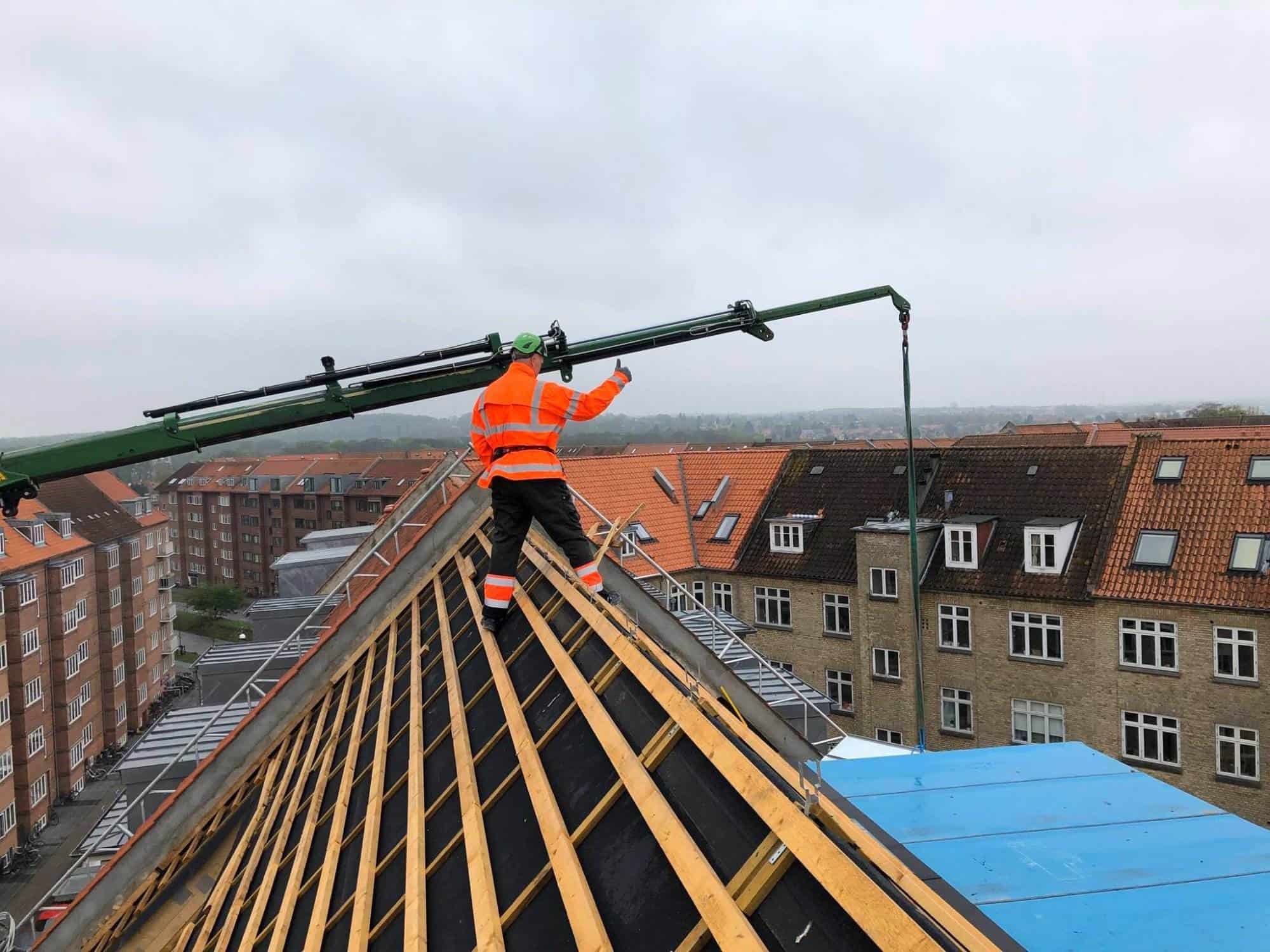 Tømrer arbejder på tagdækning med kran i bymiljø.