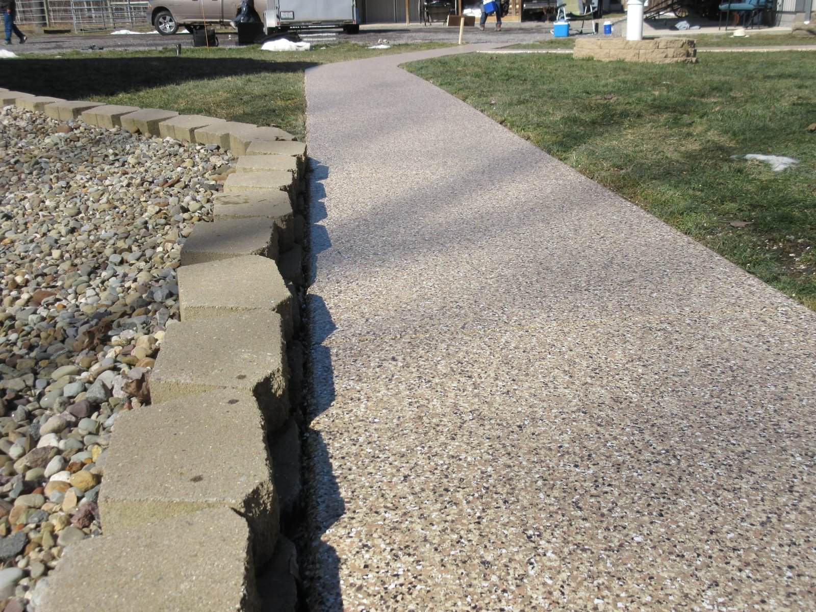 J&P Coatings made a concrete pathway that looks neat and clean. It runs through a green yard with a stone wall on one side and pebbles on the other. This shows how J&P Coatings can help make outdoor spaces look well-kept and safe to walk on. Alt text: Concrete pathway in yard improved by J&P Coatings, showing neat, safe outdoor area.