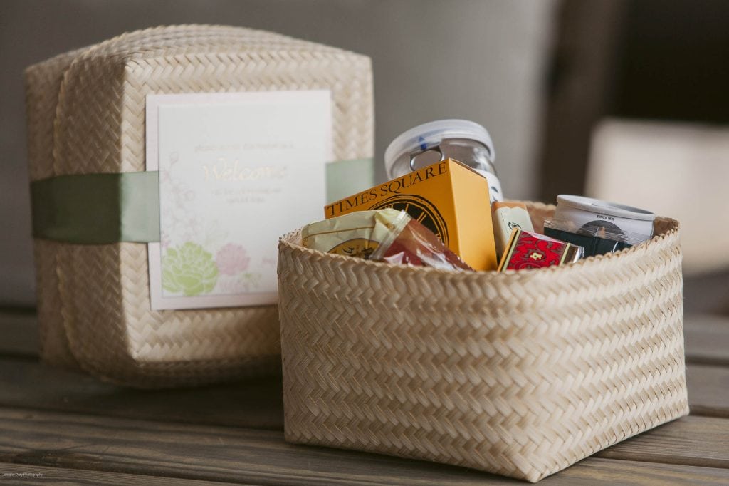 A woven basket with a lid sits open, revealing assorted snacks and small gifts inside. The lid features a pale green ribbon and a welcome card with floral designs. The basket rests on a wooden surface.