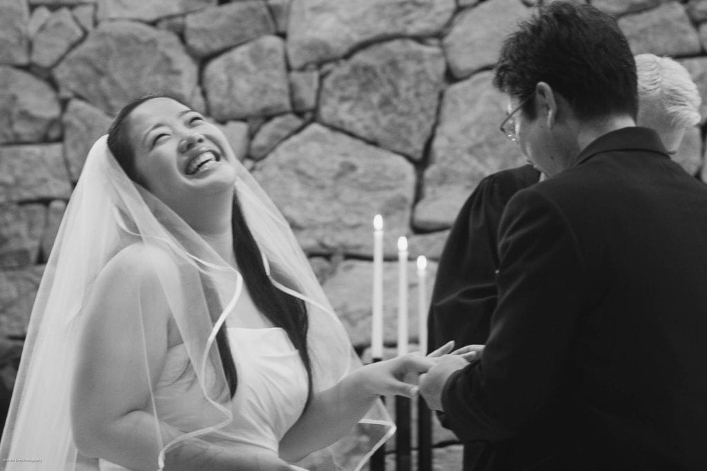 A bride in a veil laughs joyfully while holding hands with her groom during a wedding ceremony, with candles and a stone wall in the background.