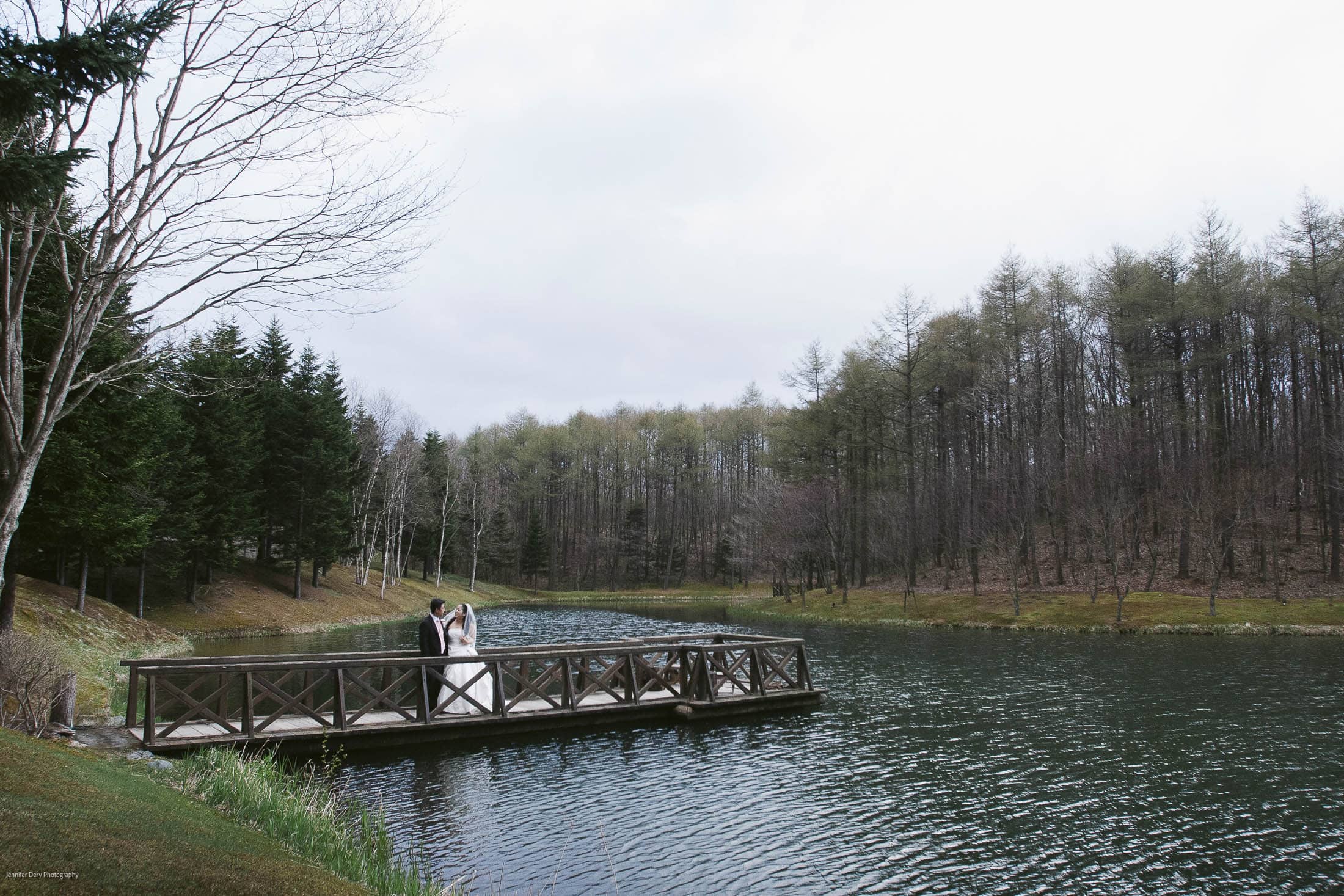 A couple in wedding attire stands on a wooden bridge over a calm lake, surrounded by trees with sparse foliage under a cloudy sky.