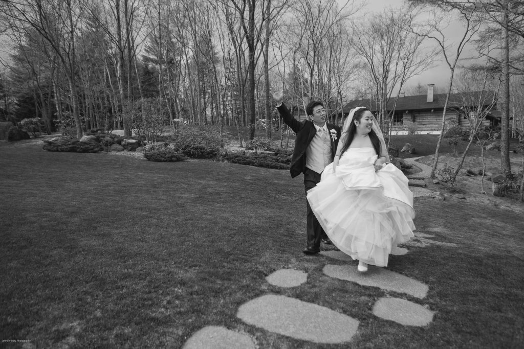A bride and groom joyfully walk on stone steps through a garden, the groom raising his arm while the bride holds up her dress. Leafless trees and a wooden cabin are visible in the background.