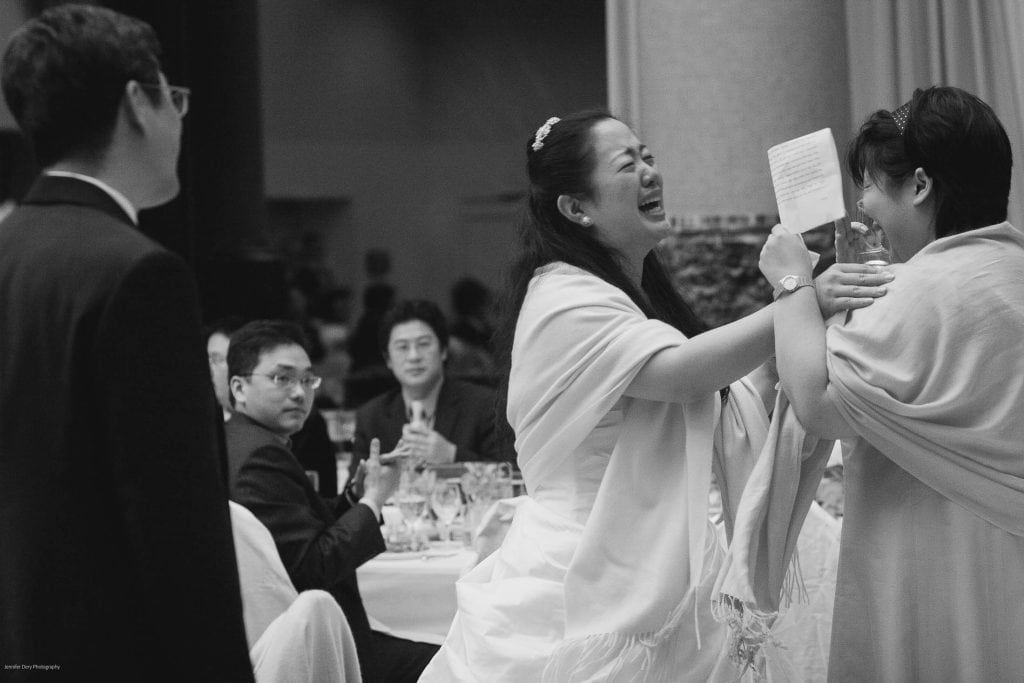 A joyful bride laughs and hugs a guest while holding a note at a wedding reception, as seated guests in suits watch and smile in the background. The scene is candid and lively, captured in black and white.