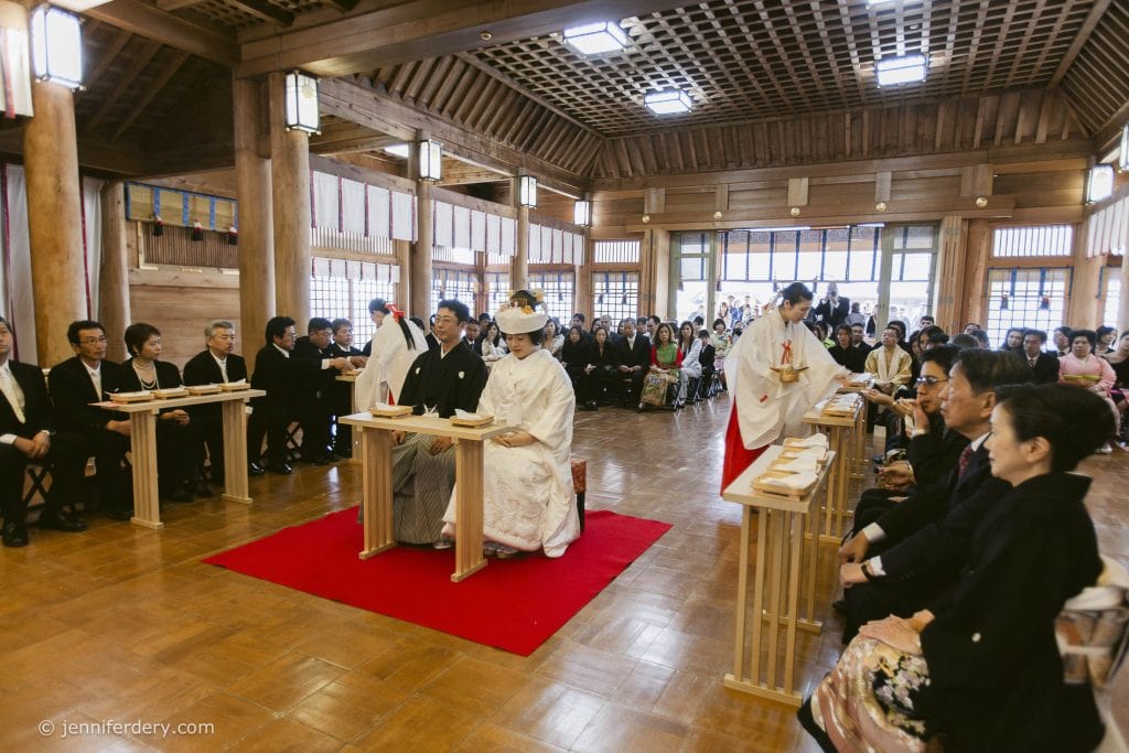 A traditional Japanese wedding ceremony in a wooden hall, with the bride and groom in white attire seated at the front, guests seated on both sides, and a priestess performing rituals.
