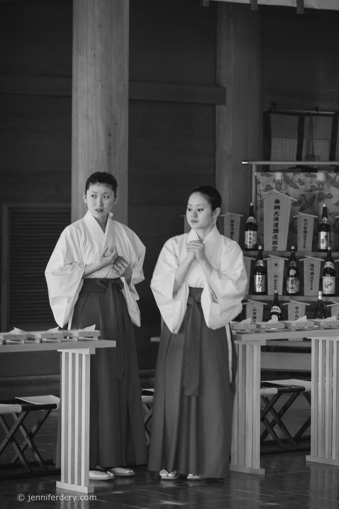 Two women dressed in traditional Japanese attire stand indoors beside wooden tables, with bottles arranged on shelves behind them. They appear to be participating in a cultural or religious ceremony. The image is in black and white.