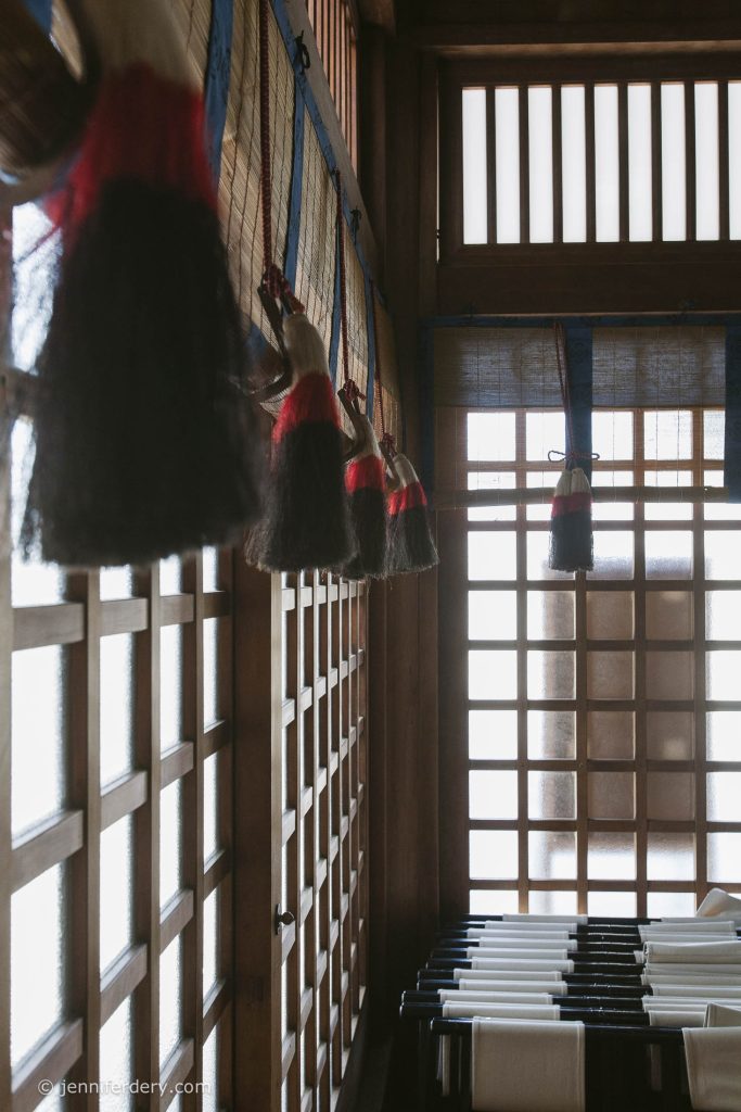 Large traditional Korean calligraphy brushes hang on a wooden wall in a sunlit room with tall, grid-patterned windows and weaving looms lined up at the bottom right.