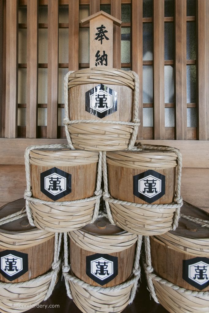 A pyramid stack of six traditional Japanese sake barrels wrapped in straw, displayed in front of a wooden wall, with a vertical wooden sign featuring Japanese characters on top.
