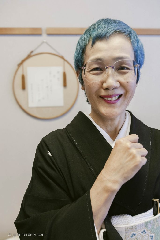 A smiling person with short blue hair and glasses wears a traditional black kimono, standing indoors with a circular wall decoration and a paper in the background.