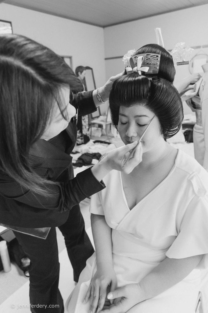 A woman in traditional Japanese attire sits with eyes closed as a makeup artist applies makeup to her face. The woman’s elaborate hairstyle is adorned with decorative hairpieces. The setting appears to be a dressing room.